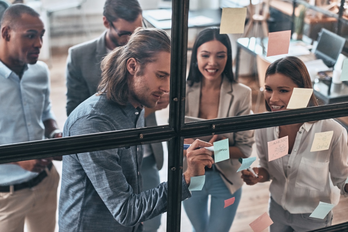 A diverse group of young professionals collaborating in a bright modern office, smiling as they write on colorful sticky notes stuck to a glass wall partition.