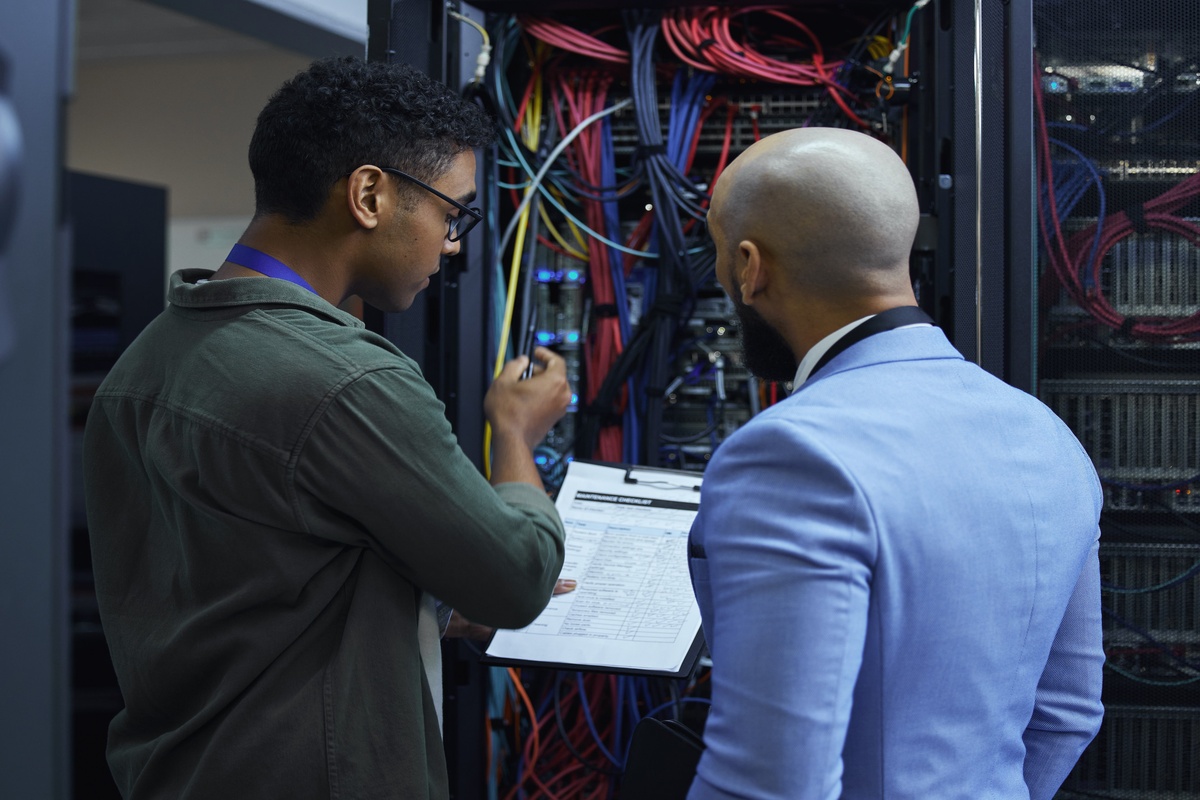 An IT technician and a manager in a light blue suit inspect a server rack filled with complex network cables while reviewing a maintenance clipboard.