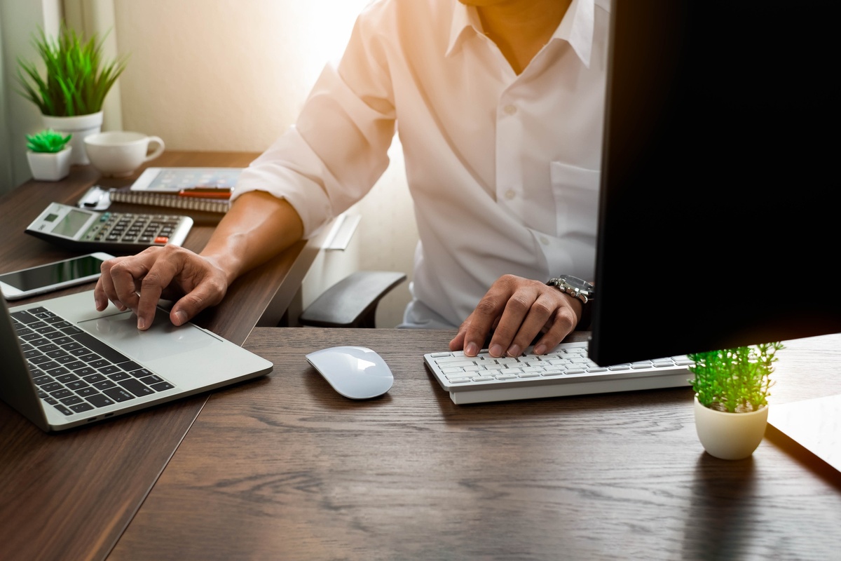 A person working at a clean wooden office desk, using a laptop trackpad with one hand and a wireless keyboard with the other.